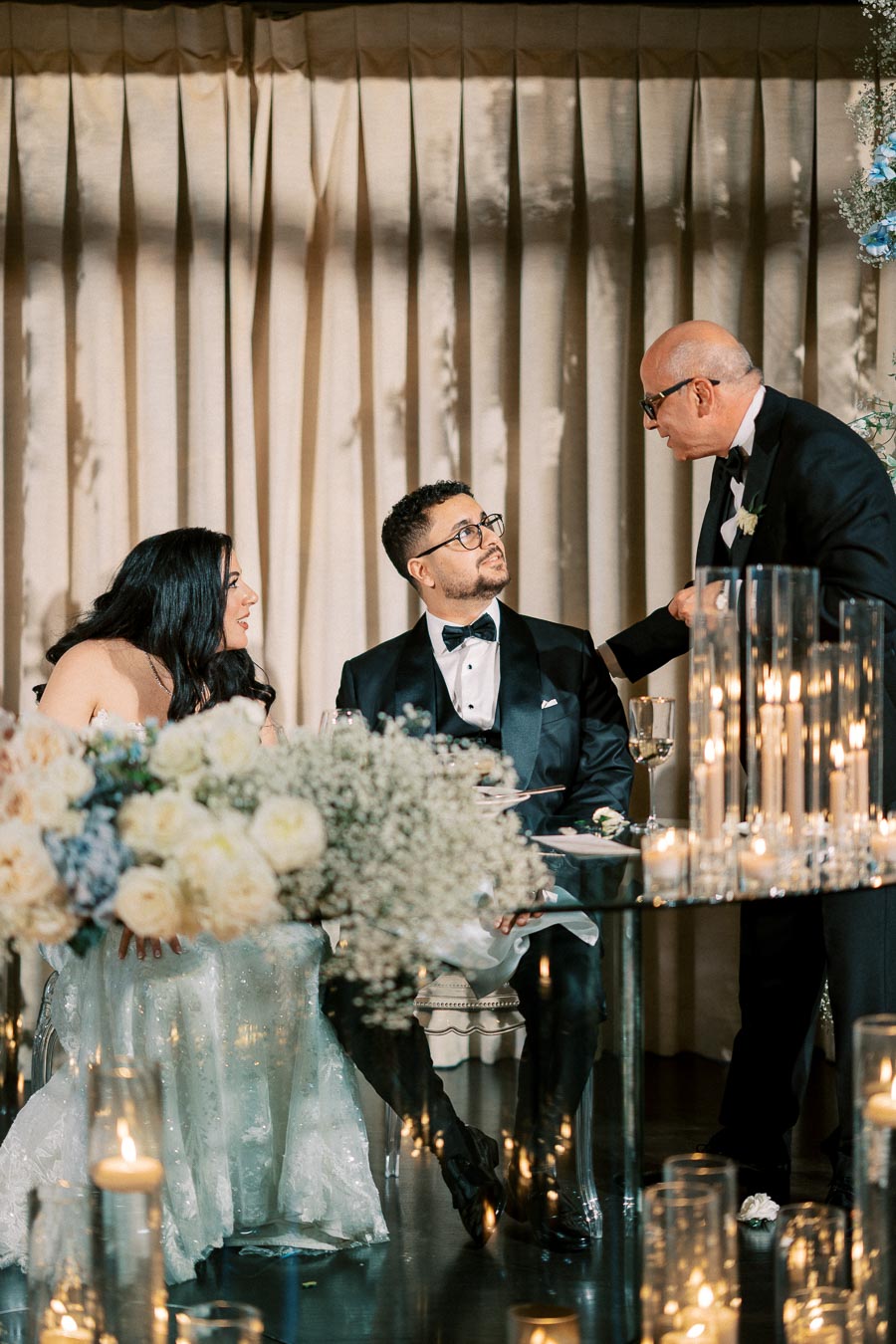 Elegant wedding reception scene with a couple seated at a table adorned with white and pastel flower arrangements. They are dressed in formal attire, listening attentively to a man in a tuxedo standing beside them. The setting is warmly lit with candles, creating a romantic ambiance.