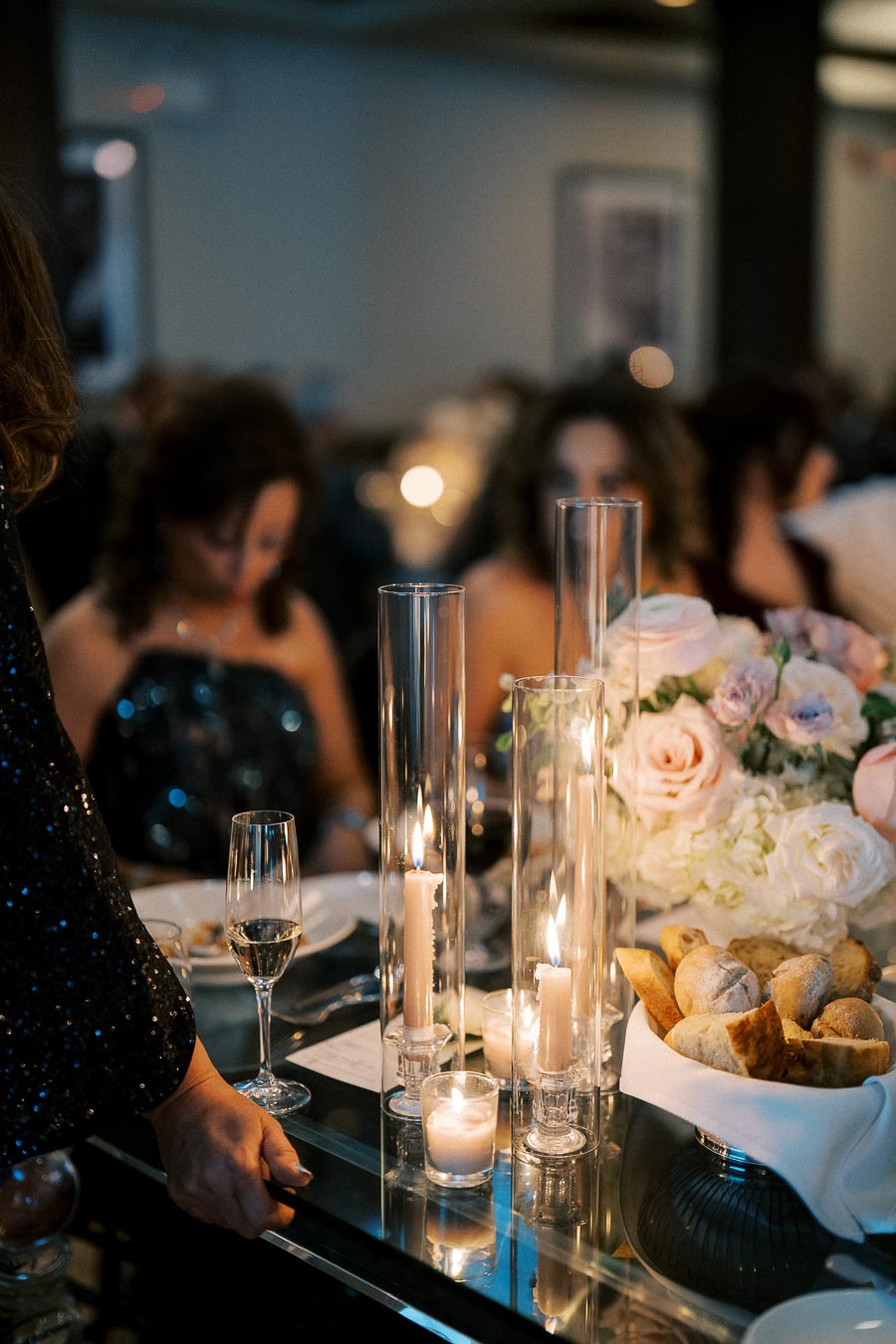 Elegant dinner table setting with lit candles, a bouquet of pastel flowers, and a basket of fresh bread rolls, creating a warm and inviting atmosphere at a formal event.
