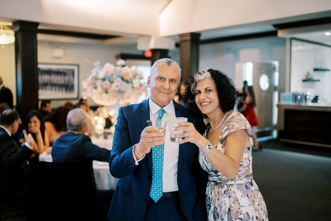 Smiling couple celebrating at a formal event, holding drinks in a beautifully decorated venue with floral centerpiece and guests in the background.