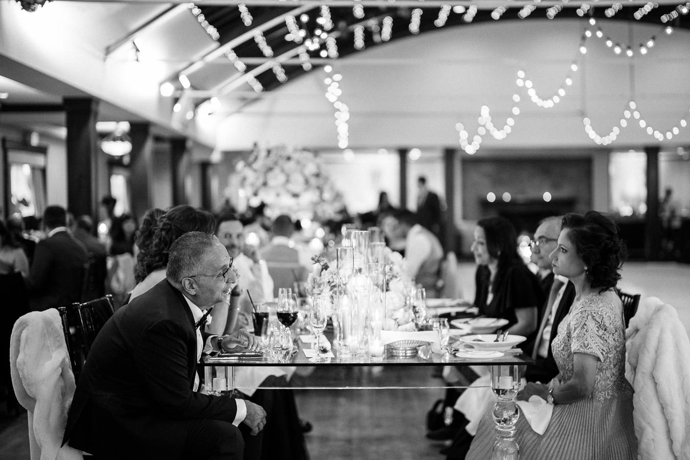 Black and white image of elegantly dressed guests seated at a formal candlelit dinner table, adorned with floral arrangements, in a decorated banquet hall.