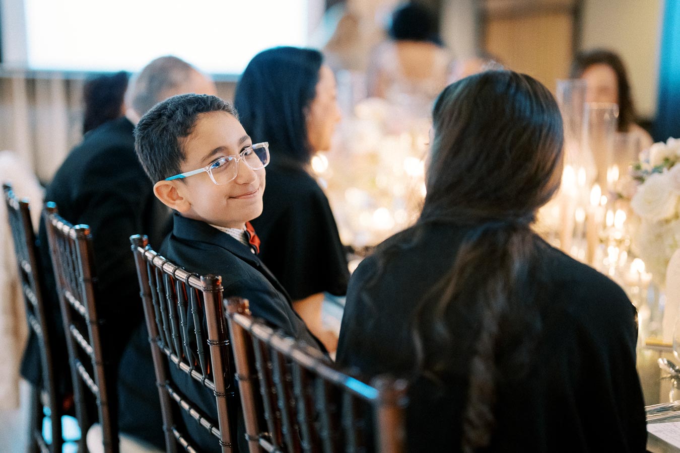 A young boy wearing glasses and a suit smiling at a formal dinner event with elegantly set tables and glowing candles.