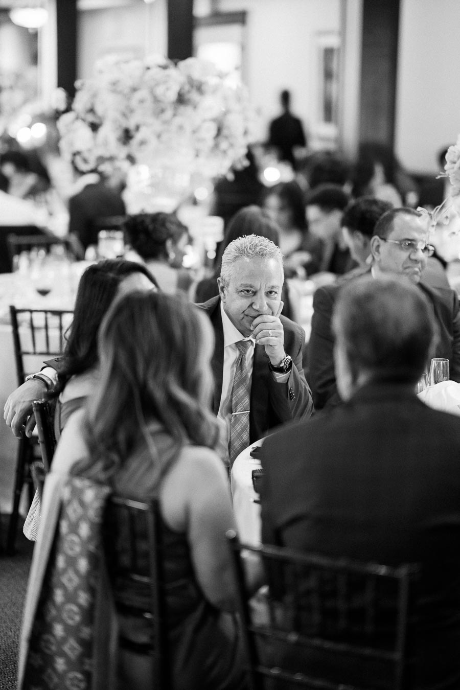 Black and white image of a social event with a group of people sitting at a table, engaged in conversation. The focus is on a man in a suit and tie, thoughtfully looking forward, while others around him interact casually. Elegant floral arrangements and ambient lighting create a sophisticated atmosphere.