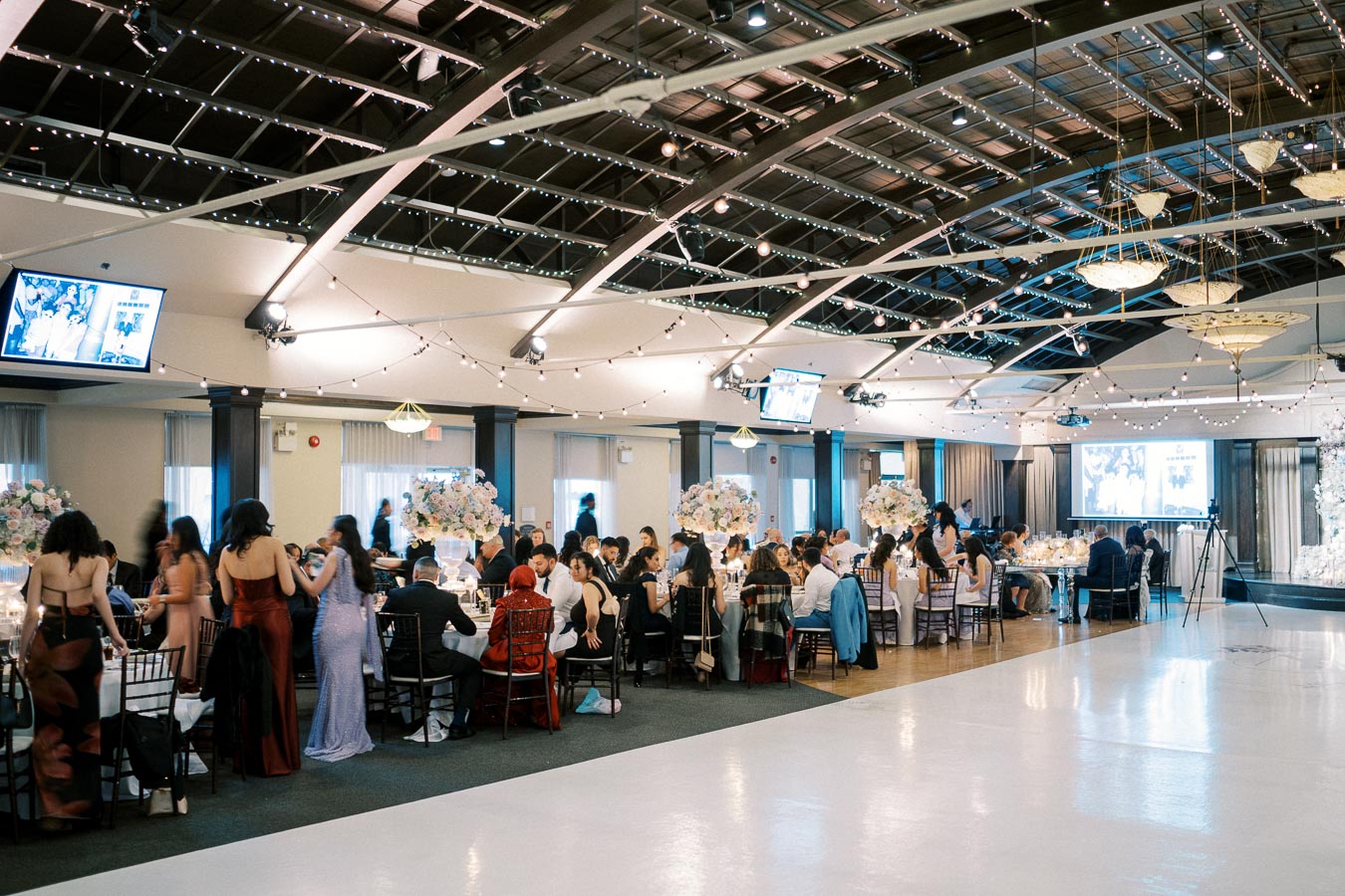 Elegant wedding reception in a spacious hall featuring decorated tables with floral centerpieces, guests in formal attire, and ambient lighting from overhead string lights and chandeliers.