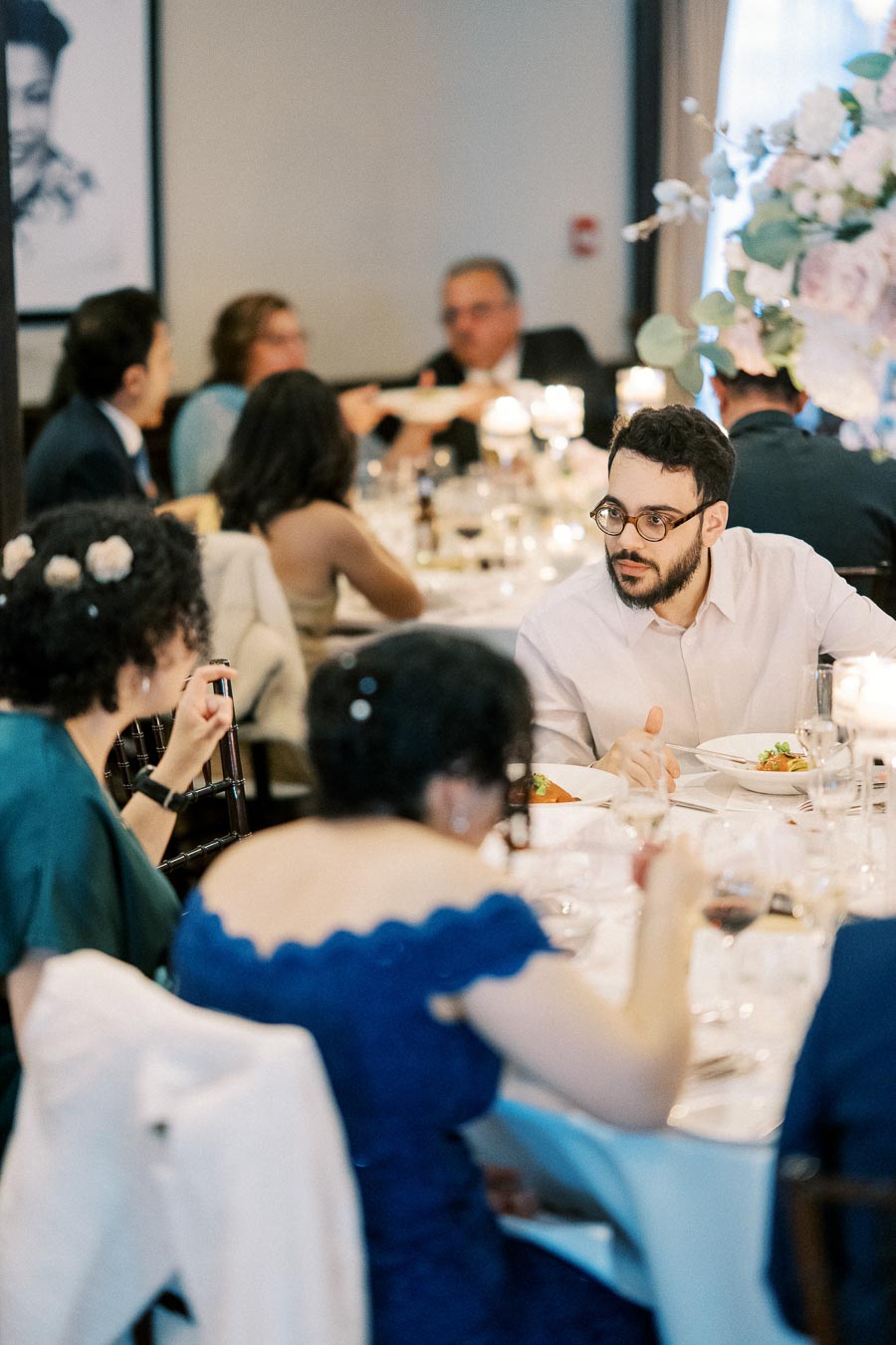 A group of people seated at an elegant, candle-lit dinner event, engaging in conversation around a beautifully set table with floral centerpieces and gourmet dishes.