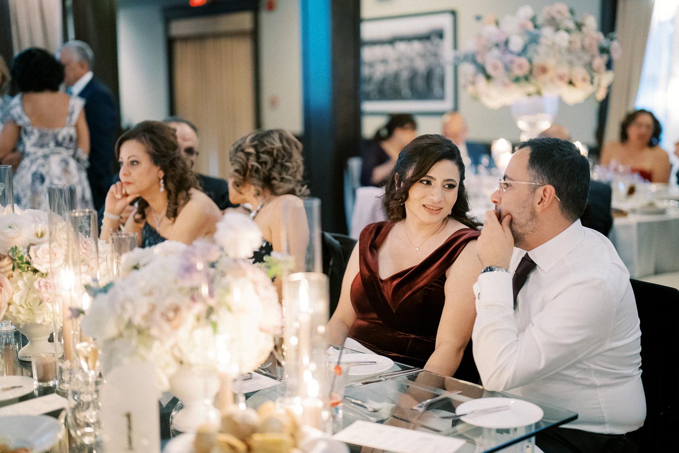 Elegant wedding reception with formally dressed guests seated at a beautifully decorated table with floral centerpieces, candles, and ambient lighting.