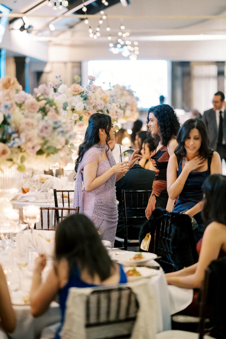 Elegant wedding reception with guests socializing in a beautifully decorated hall, featuring floral centerpieces and ambient string lighting.