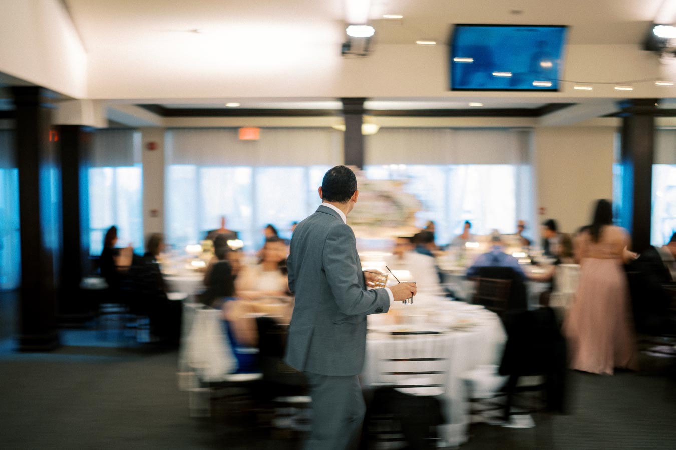 A man in a suit walking through a blurred banquet hall, holding papers and a pen, with guests seated at round tables in the background.