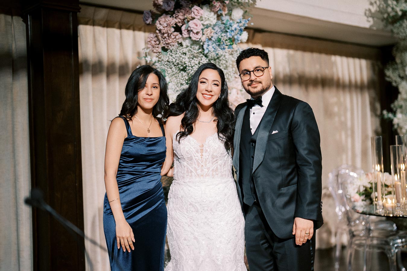 Three people posing at a formal event, dressed elegantly in a wedding setting with floral decorations in the background.