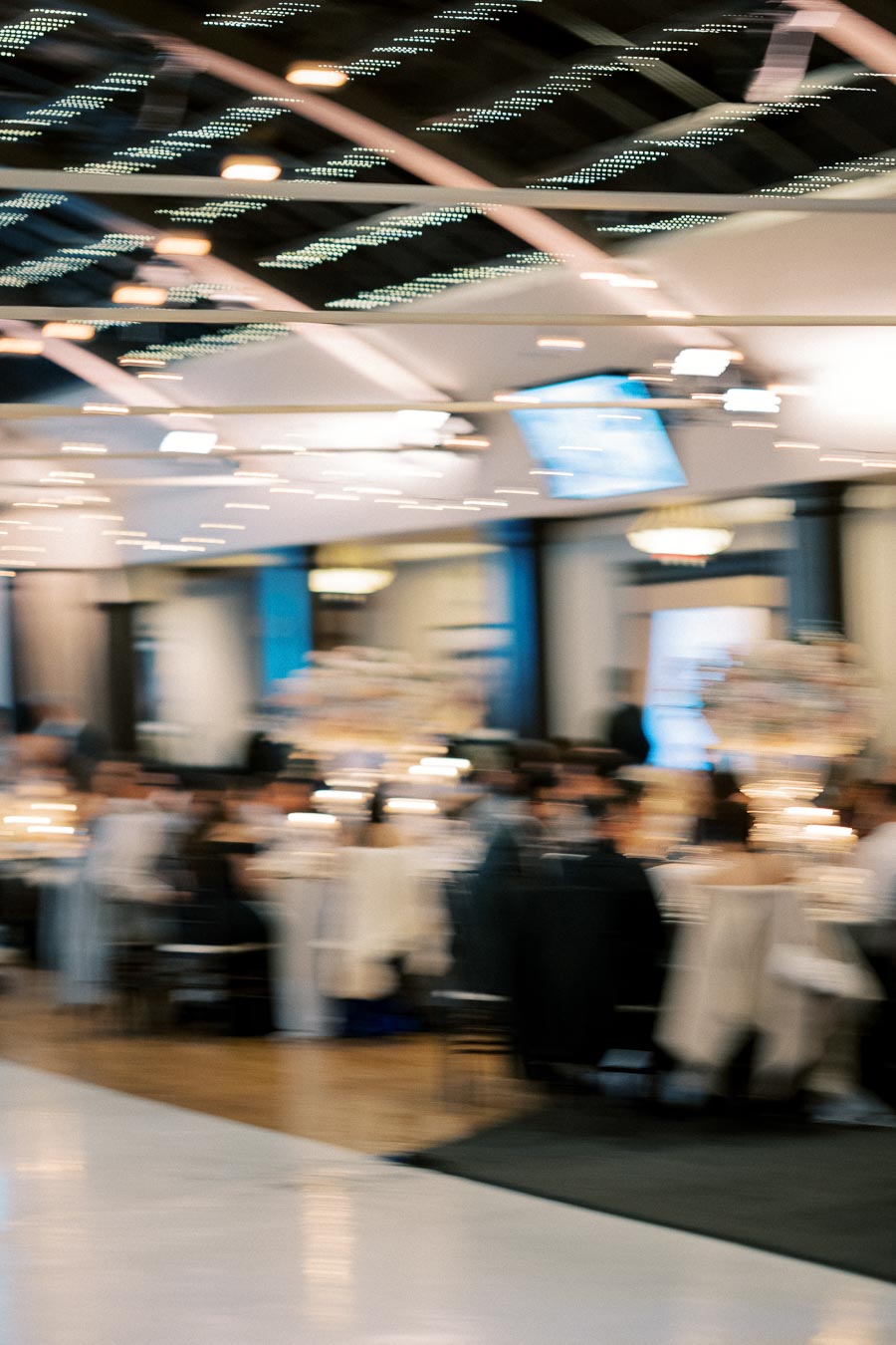 Blurred image of an elegant indoor event space with guests seated at tables, featuring softly glowing lights and decorative elements hanging from the ceiling.