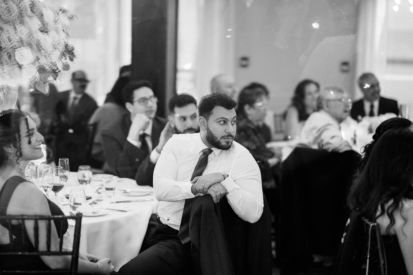 Black and white image of people seated at a formal event, attentively watching a presentation. The foreground shows a man in a suit with his jacket draped over a chair, surrounded by other guests at elegantly set tables.