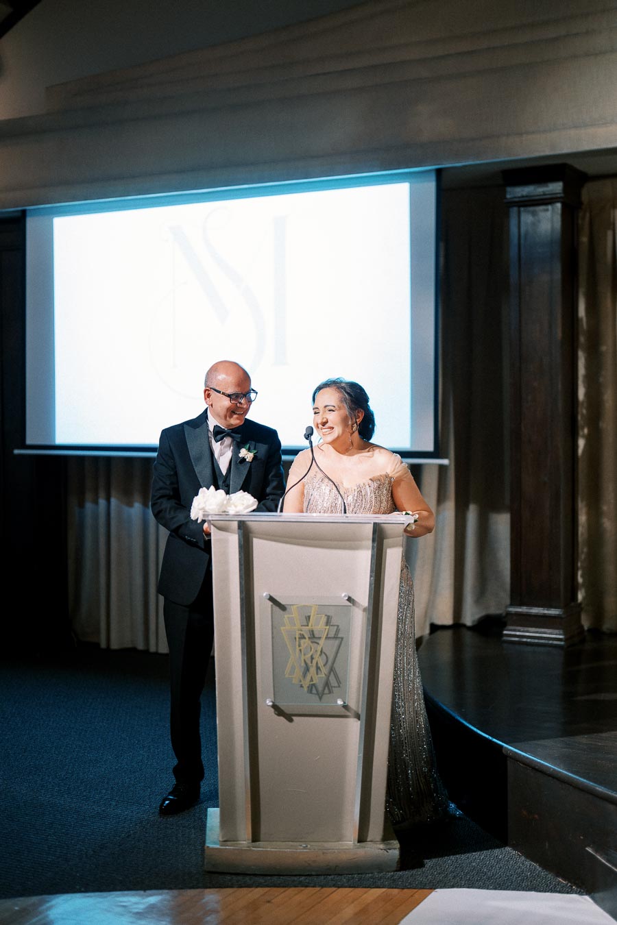 A couple dressed in formal attire, speaking at a podium during a special event, with a projected logo in the background.