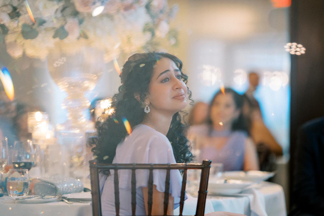 A person with curly hair and earrings sits at an elegant dinner table, surrounded by soft lighting and a blurred background, conveying a serene and contemplative atmosphere.
