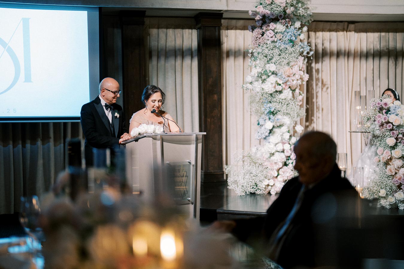 Elegant wedding reception with a man and woman at a podium, speaking into a microphone, surrounded by floral decorations and soft candlelight ambiance.