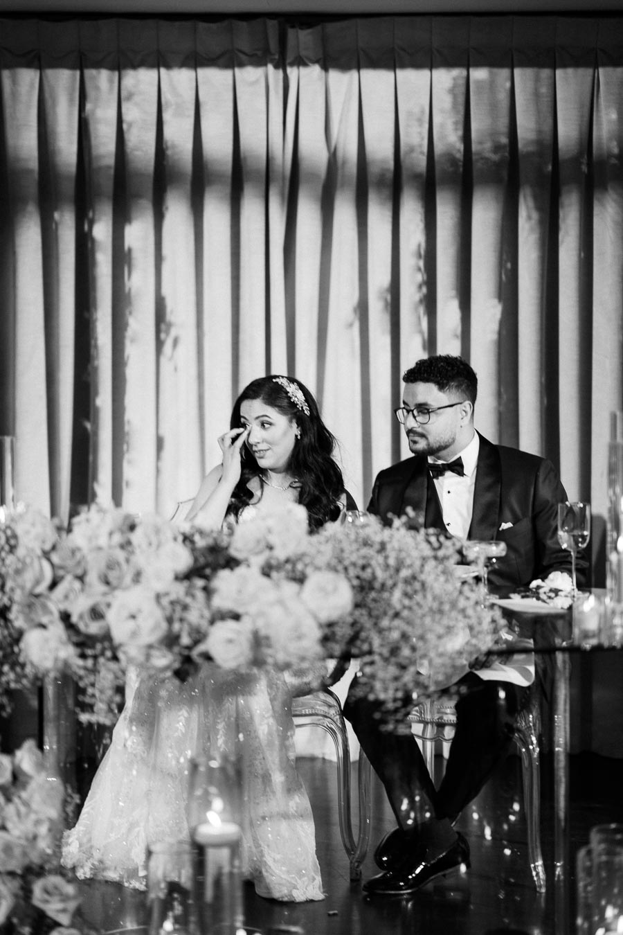 Black and white image of a wedding couple seated at their reception, surrounded by floral arrangements. The bride, wearing an elegant gown and headpiece, appears emotional, while the groom, in a tuxedo and glasses, looks on with a thoughtful expression.