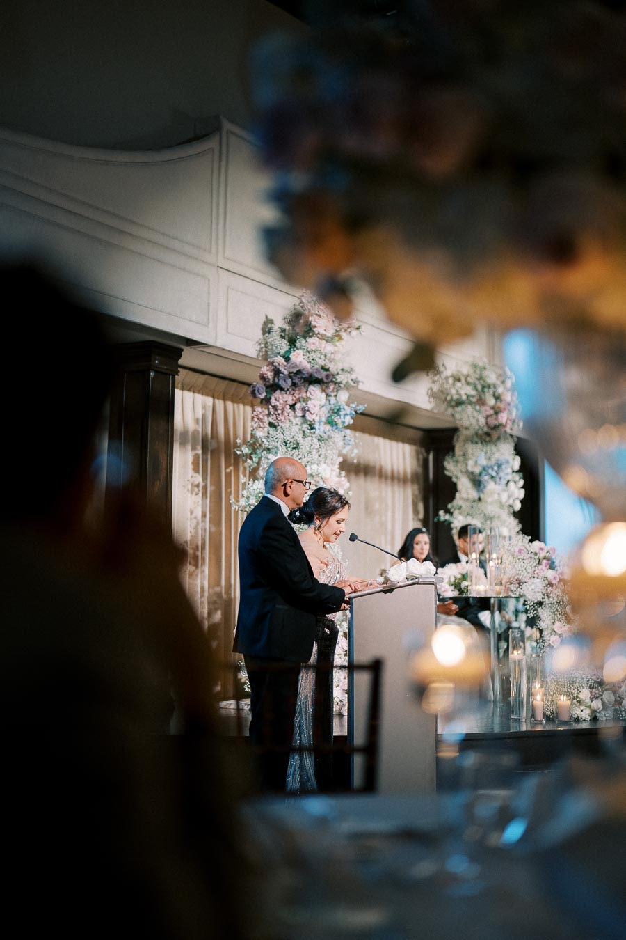 Elegant wedding reception speech with couple at podium, surrounded by floral arrangements, soft lighting, and guests seated in background.