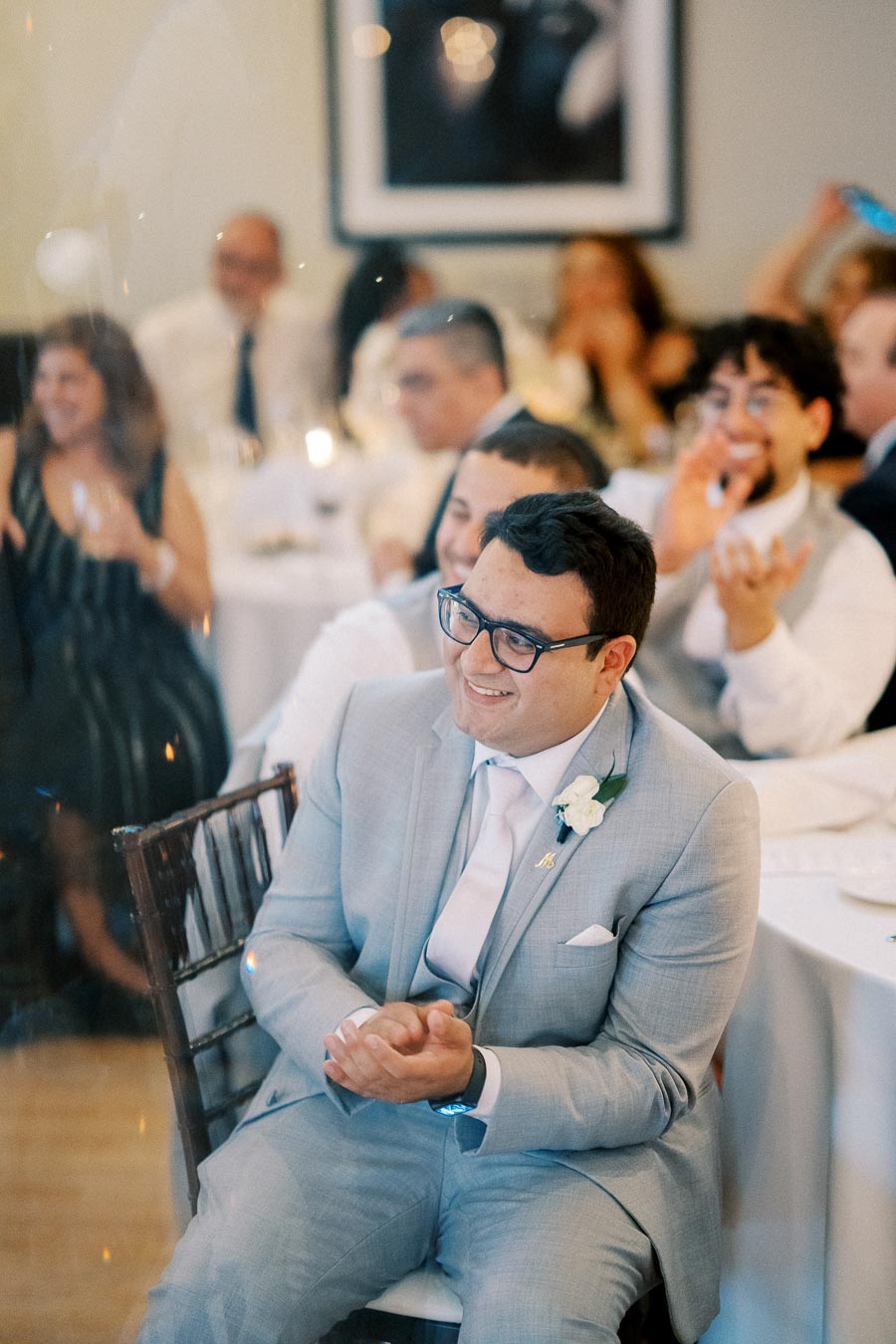 A man in a light gray suit with a floral boutonniere claps and smiles while seated at a wedding reception, surrounded by other guests in formal attire.