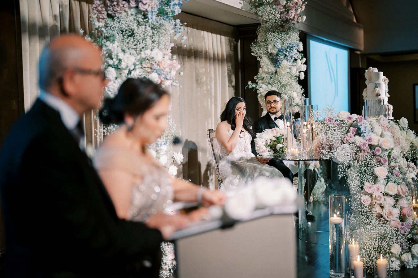 Bride and groom sitting at a beautifully decorated wedding reception table, surrounded by elegant floral arrangements and candles, as they listen emotionally to speeches.