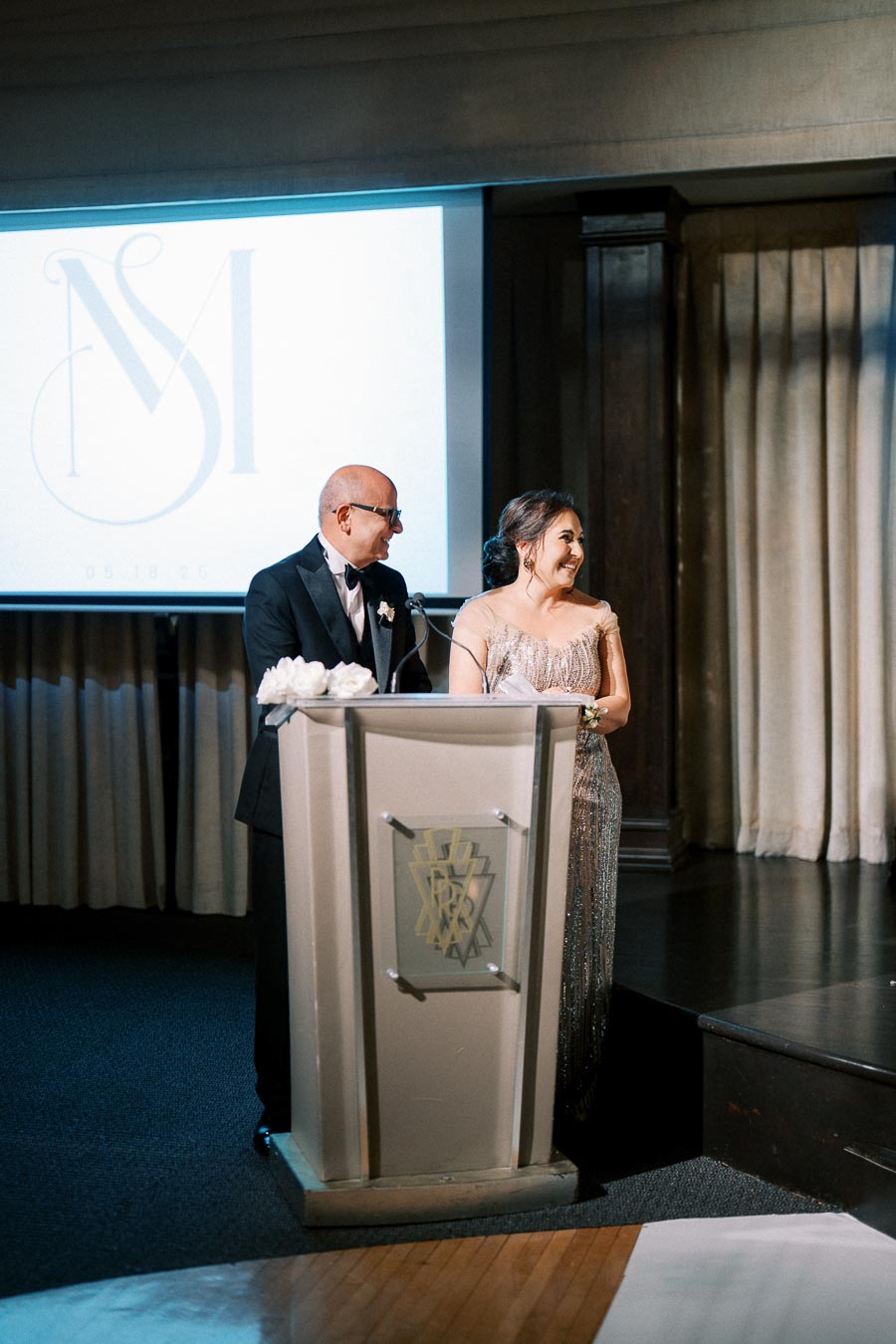 Couple standing at a podium during an elegant event, with a logo projected on a screen behind them. They are dressed in formal attire, smiling and engaging with the audience.