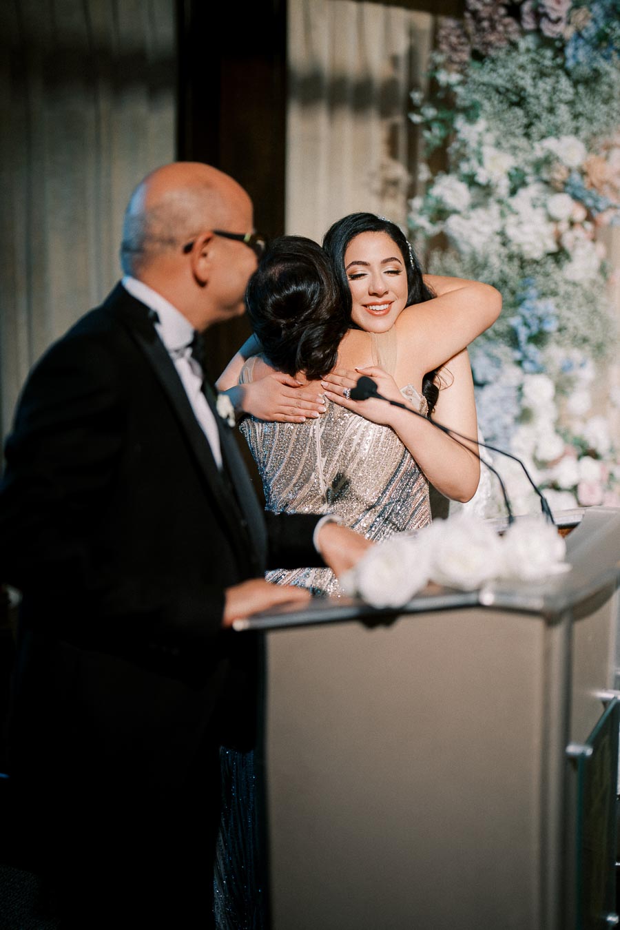 A woman in an elegant dress hugs another person warmly at a formal event, with a man in a suit standing nearby at a podium decorated with white flowers. A backdrop of flowers enhances the celebratory atmosphere.