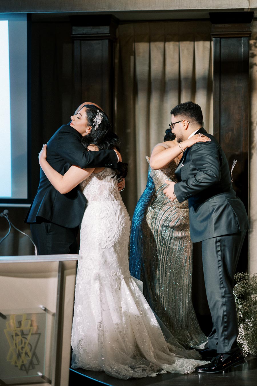 Two couples embrace in a heartfelt hug during a formal wedding ceremony, with the bride in an elegant white gown and the groom in a sleek suit.