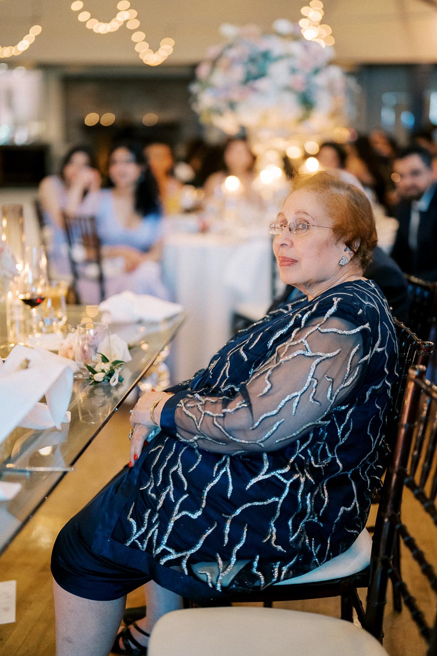 Elderly woman in elegant blue dress sitting at a wedding reception, surrounded by beautifully decorated tables and warm lighting.