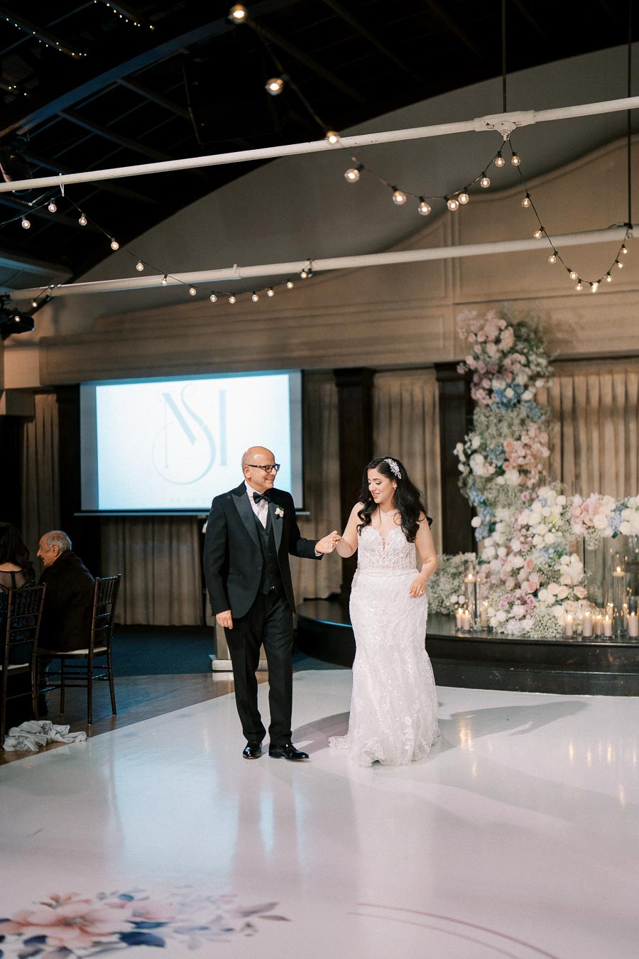 A bride in a white gown dances with a man in a tuxedo at a wedding reception, with elegant floral decorations and soft lighting enhancing the romantic atmosphere.