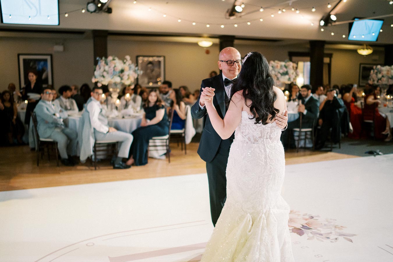 A bride in a white wedding dress dances with an older man in formal attire at a wedding reception, surrounded by elegantly dressed guests seated at decorated tables under soft, ambient lighting.