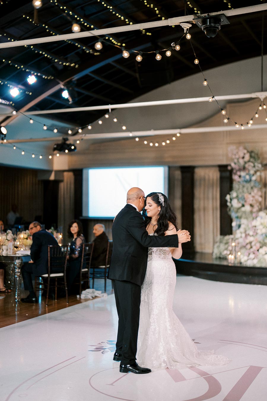 A bride and her father sharing a heartfelt dance on a beautifully decorated wedding dance floor, with elegant string lights and floral arrangements in the background.