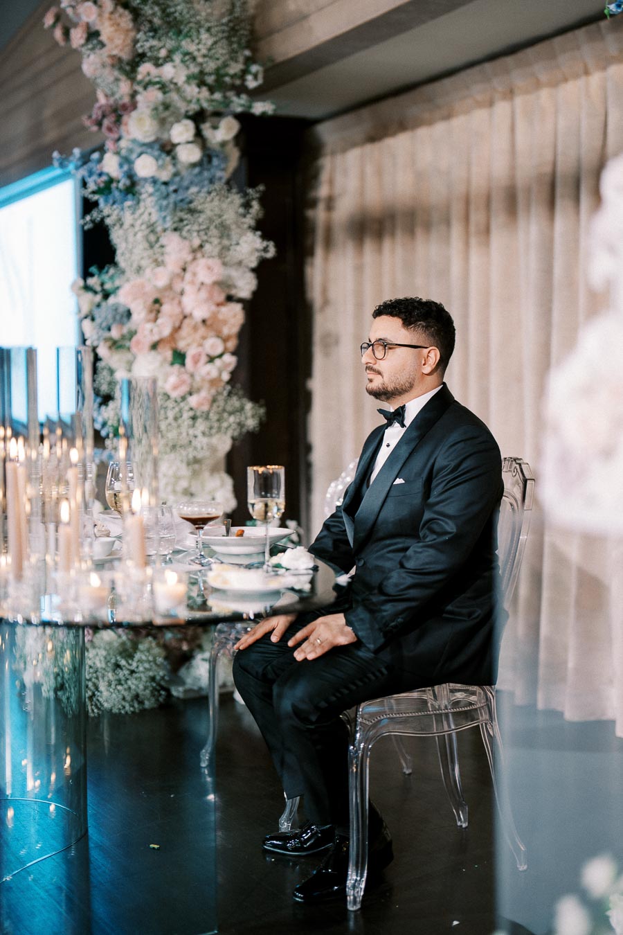 A man in a formal black tuxedo sits at an elegant table decorated with candles and floral arrangements during a wedding reception. The atmosphere exudes sophistication and celebration.