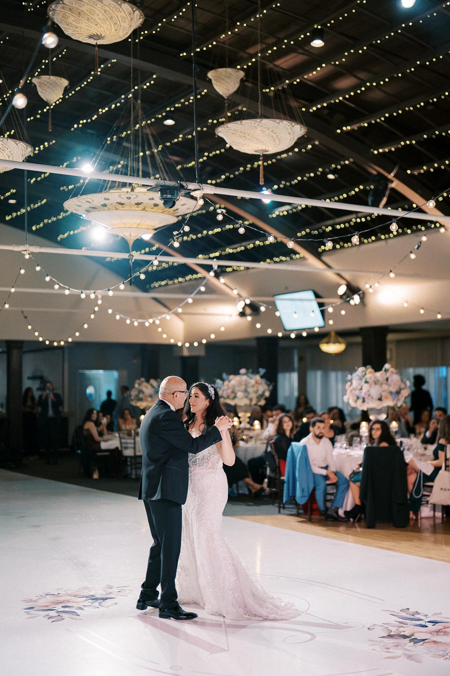 Father and bride dancing during a wedding reception under decorative string lights, surrounded by seated guests with elegant floral arrangements.