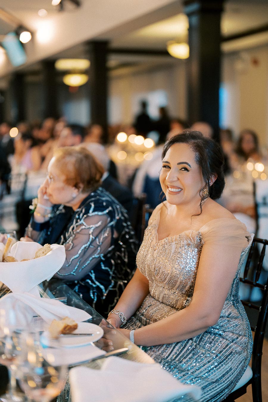 Smiling woman in a sparkling evening gown sitting at a banquet table during a formal event, with other guests visible in the background.