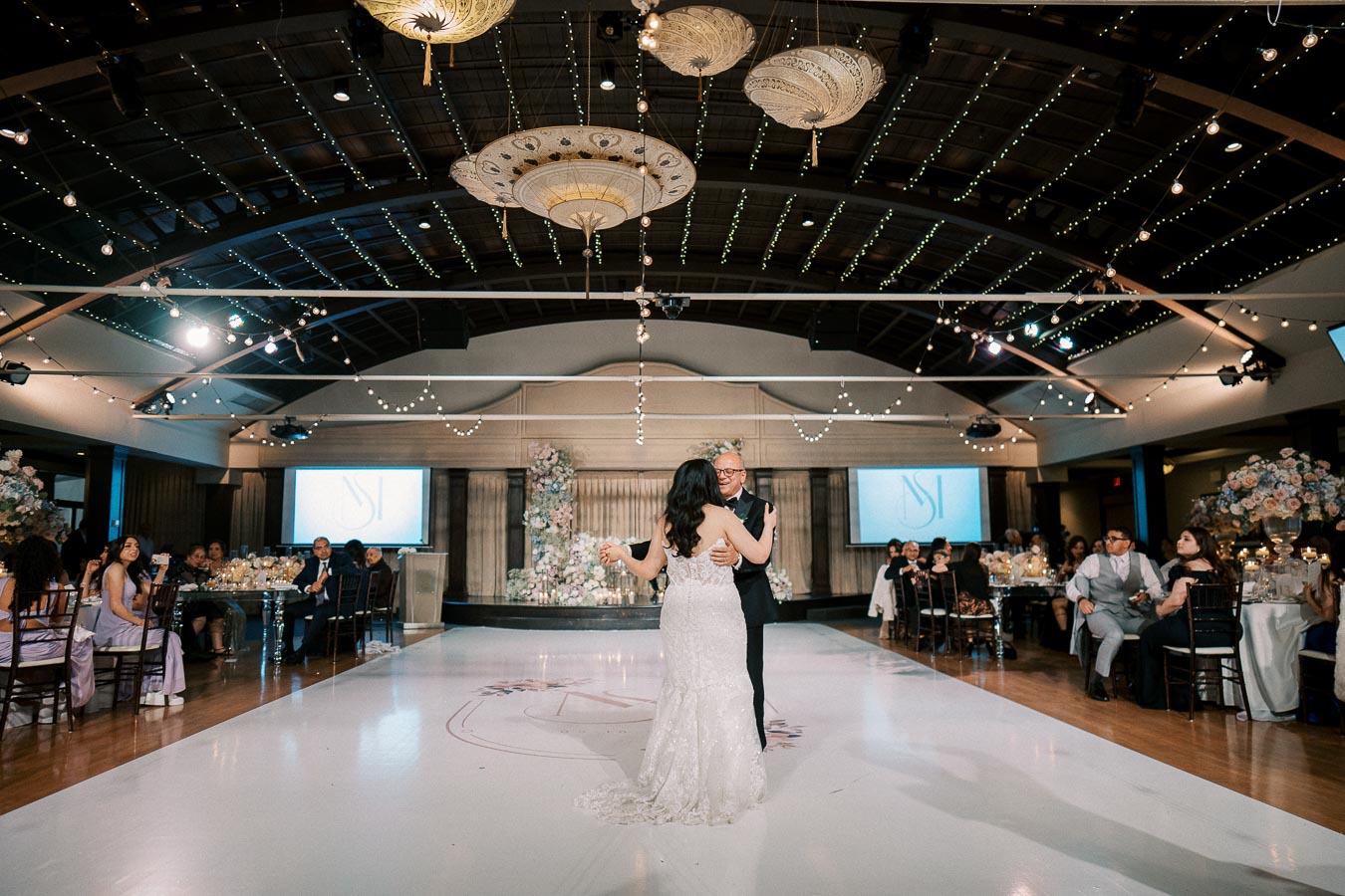 A couple shares their first dance on a polished dance floor in an elegantly decorated wedding venue, adorned with floral arrangements and string lights, surrounded by seated guests.