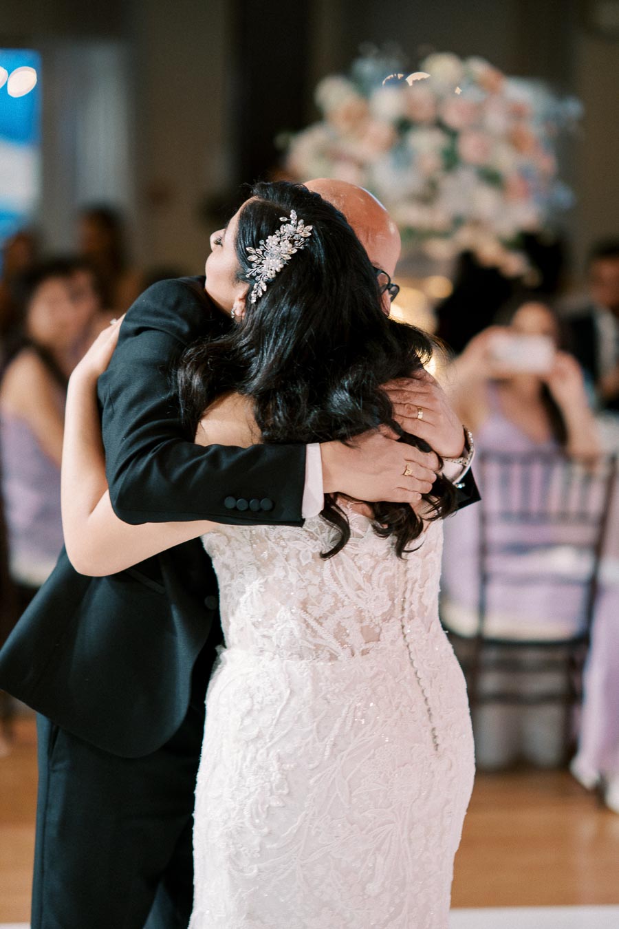 Wedding couple embrace during first dance at reception, bride in elegant white lace dress with decorative hair accessory, surrounded by guests in a softly lit venue.