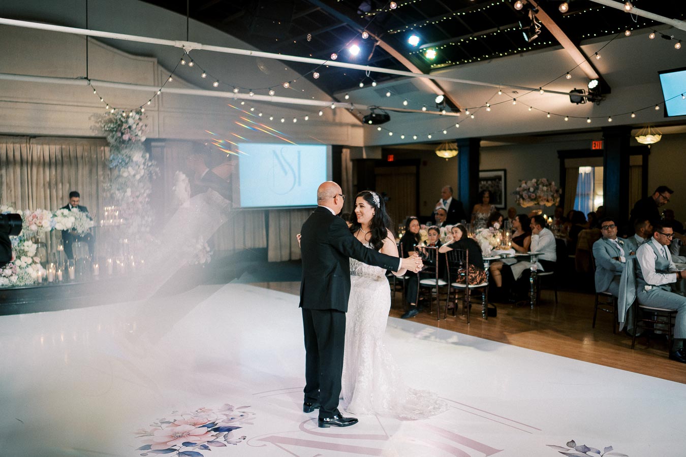 A father and daughter share a joyful dance at a wedding reception, surrounded by elegantly decorated tables and seated guests in a warmly lit venue.
