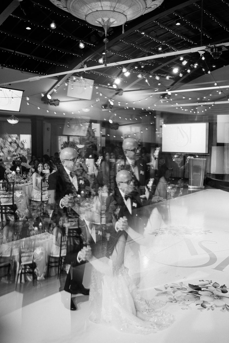 Black and white photo of a wedding reception with a double exposure effect, showing a couple dancing on a decorated dance floor adorned with floral patterns, amidst a ceiling of string lights and elegantly dressed guests seated at tables.