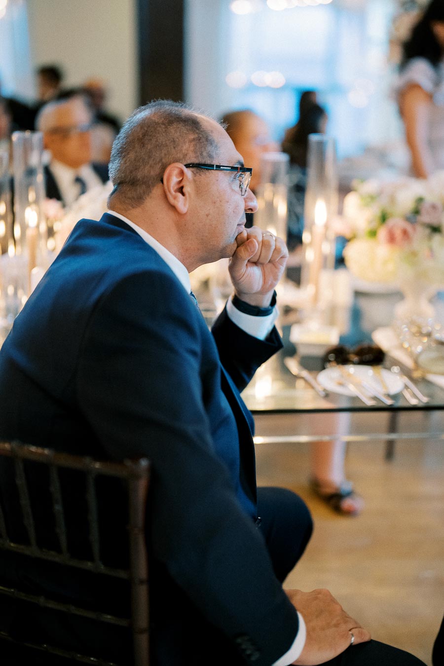 A thoughtful man in a suit sitting at a formal event table, surrounded by elegant decor and soft candlelight.