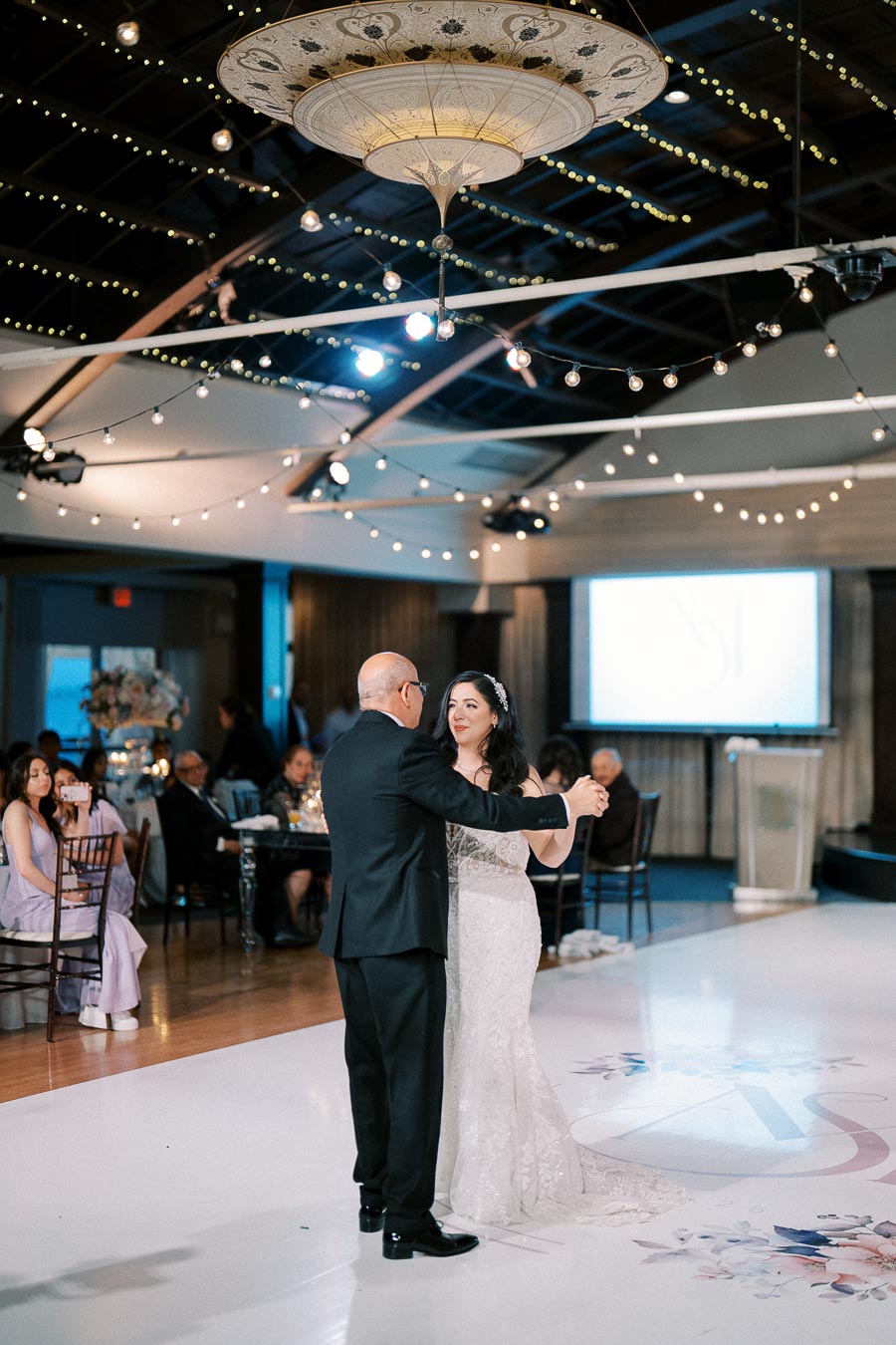A bride and her father share a dance at a wedding reception, surrounded by elegantly dressed guests and soft ambient lighting with string lights hanging from the ceiling of the venue.
