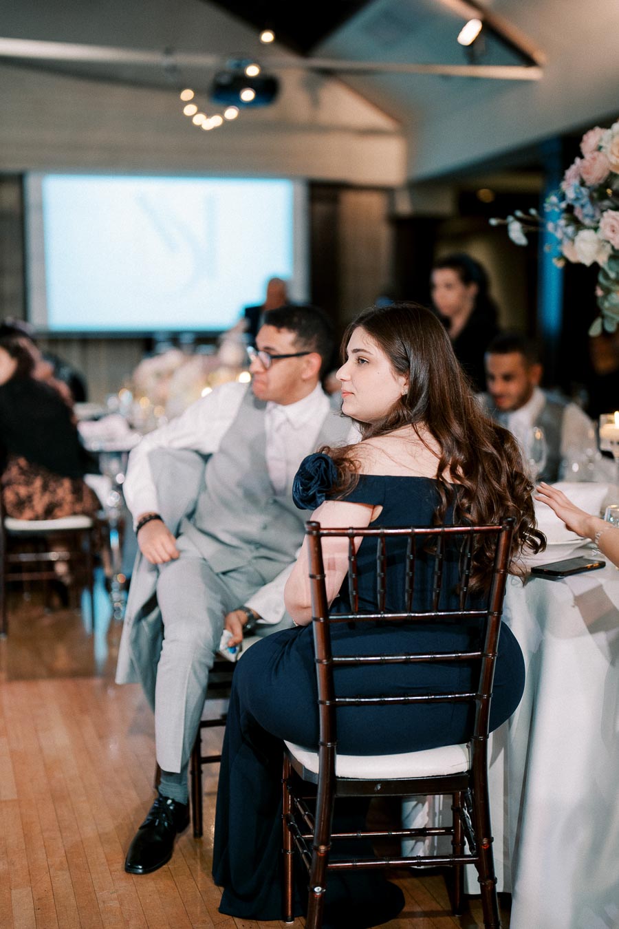 Alt text: Elegantly dressed guests sitting at a formal event, with a presentation screen in the background and floral decorations on the table.