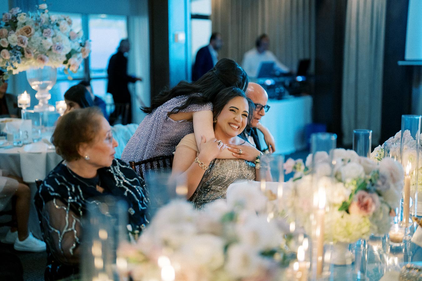 A joyful moment at a wedding reception, featuring guests embracing and smiling amidst elegant floral arrangements and candlelit tables, with a DJ setup in the background.