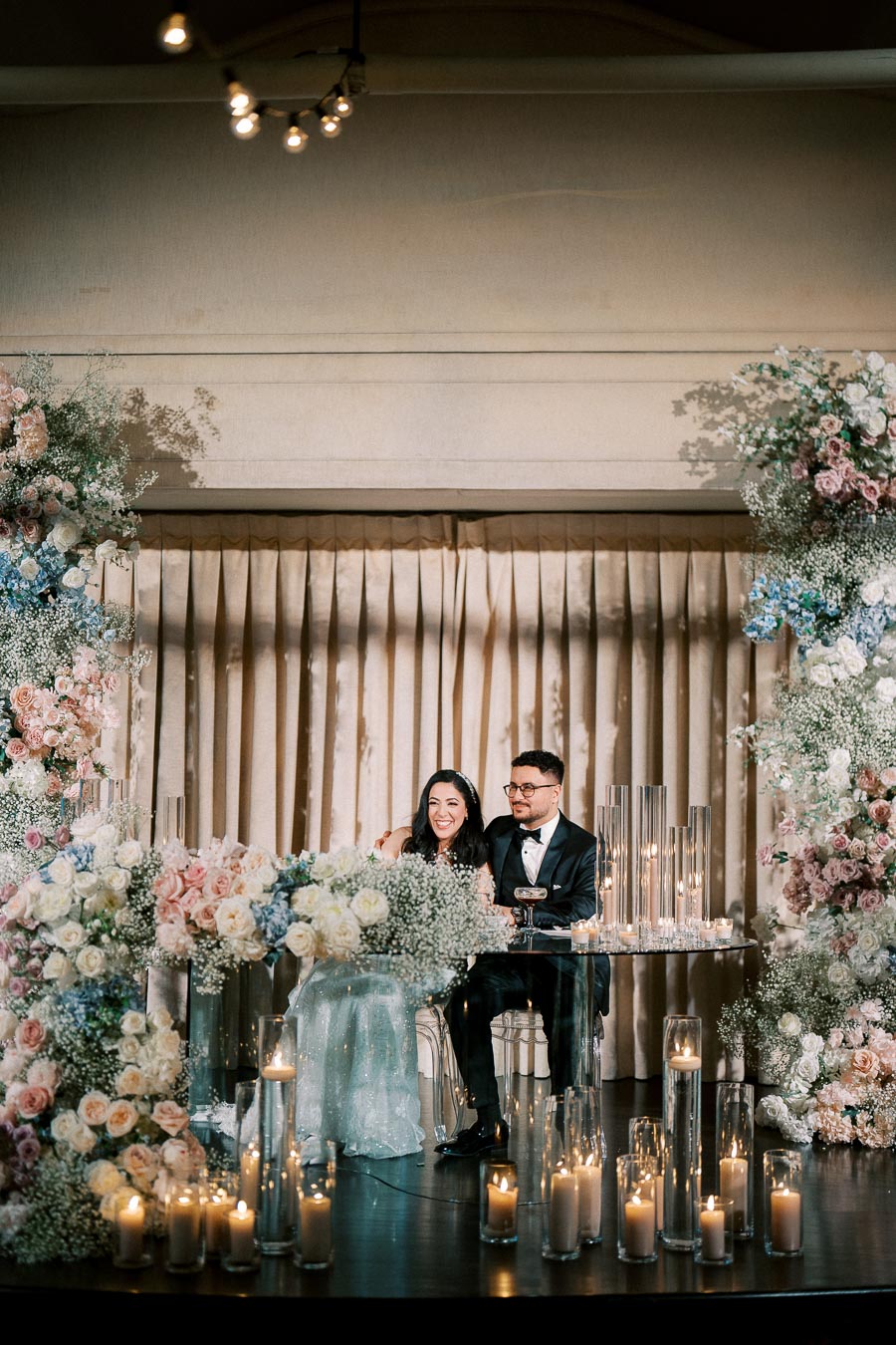 A happy couple sitting at an elegantly decorated wedding reception table, adorned with a lush arrangement of pink, white, and blue flowers, surrounded by candles in tall glass holders, creating a romantic and enchanting atmosphere.