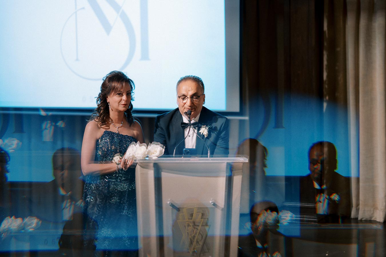 Two elegantly dressed individuals at a podium speaking into a microphone, with a projected logo on a screen in the background during a formal event.