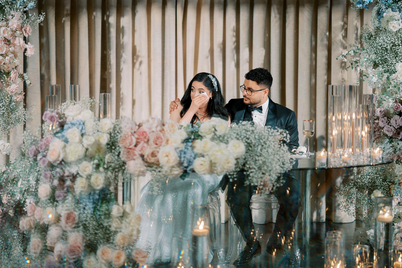 A bride and groom sitting together at an elegantly decorated wedding reception table. The bride wipes a tear with a tissue, surrounded by lush floral arrangements and candlelight. The groom looks on with a caring expression, providing a comforting presence.