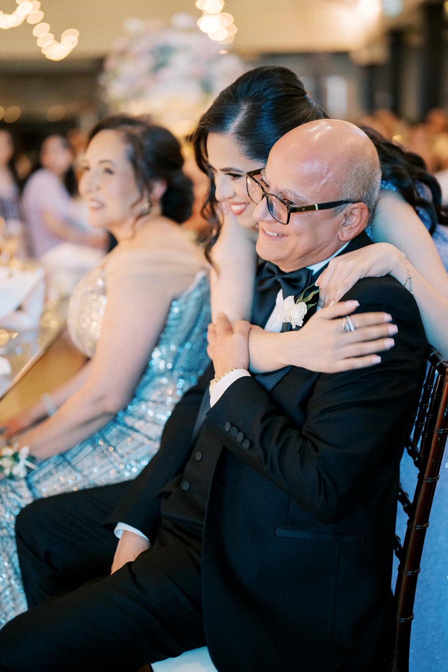 A joyful moment at a wedding reception with a woman in an elegant dress embracing an older man in a tuxedo, both smiling and surrounded by soft lighting and blurred guests in the background.