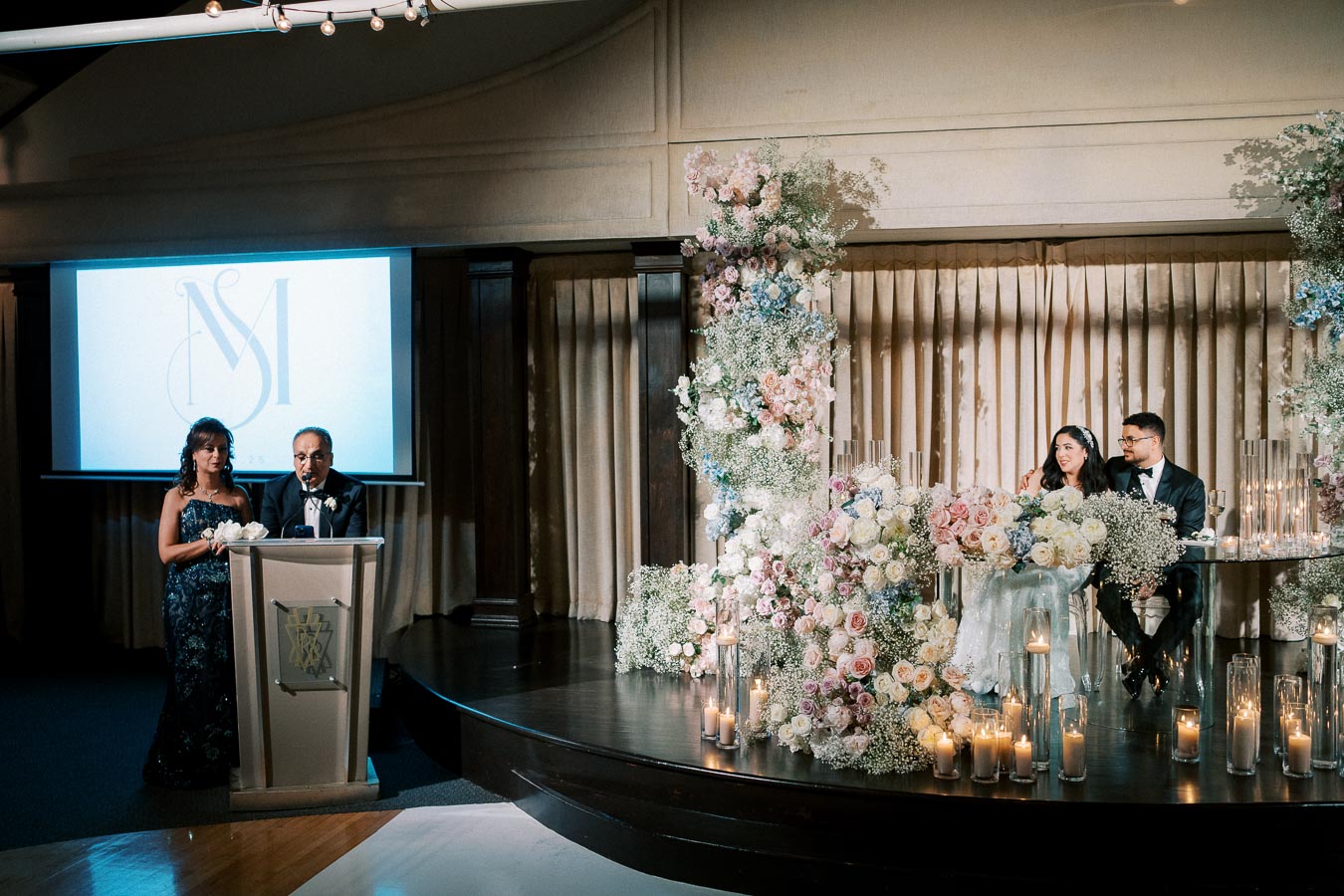 Elegant wedding reception with a couple sitting at a beautifully decorated table adorned with flowers and candles; speakers giving a speech at a podium; ambient lighting enhances the romantic atmosphere.