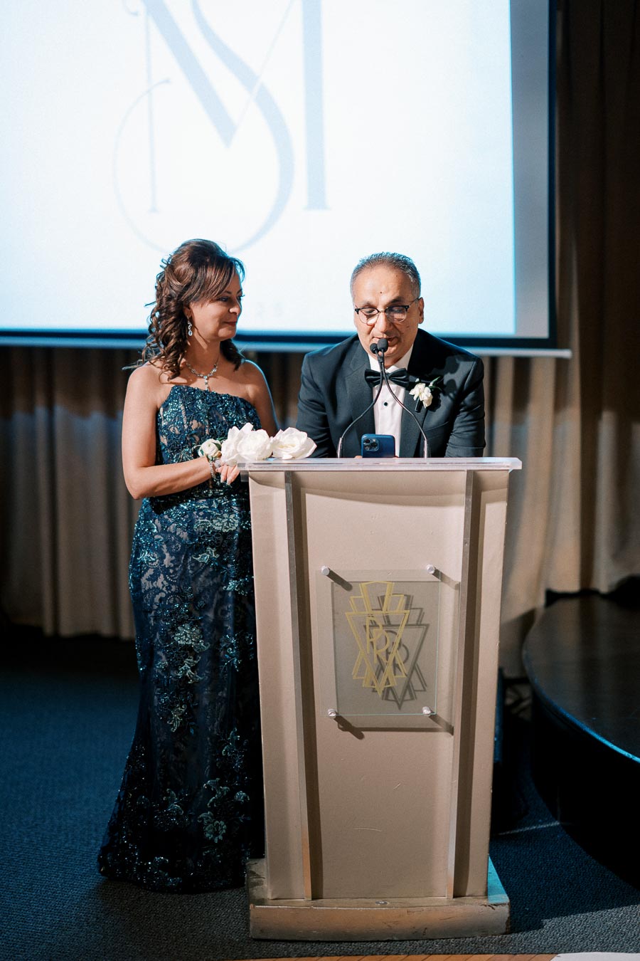 A man and woman in formal attire stand at a podium giving a speech at an event, with a screen featuring a stylized logo in the background.
