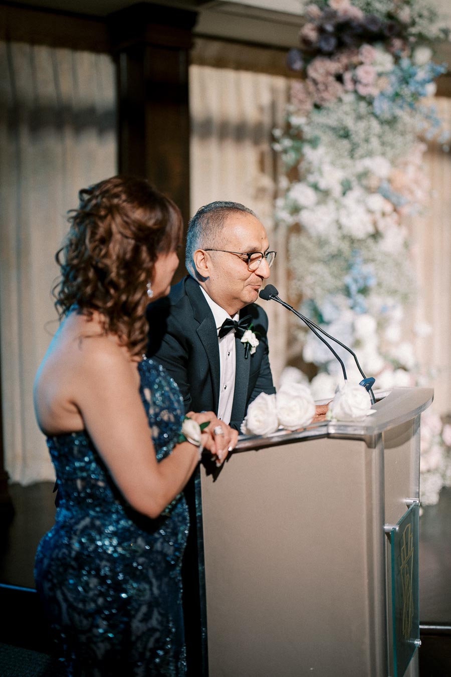 Formal event speakers at a podium, elegantly dressed in evening attire, delivering a speech with a floral backdrop.