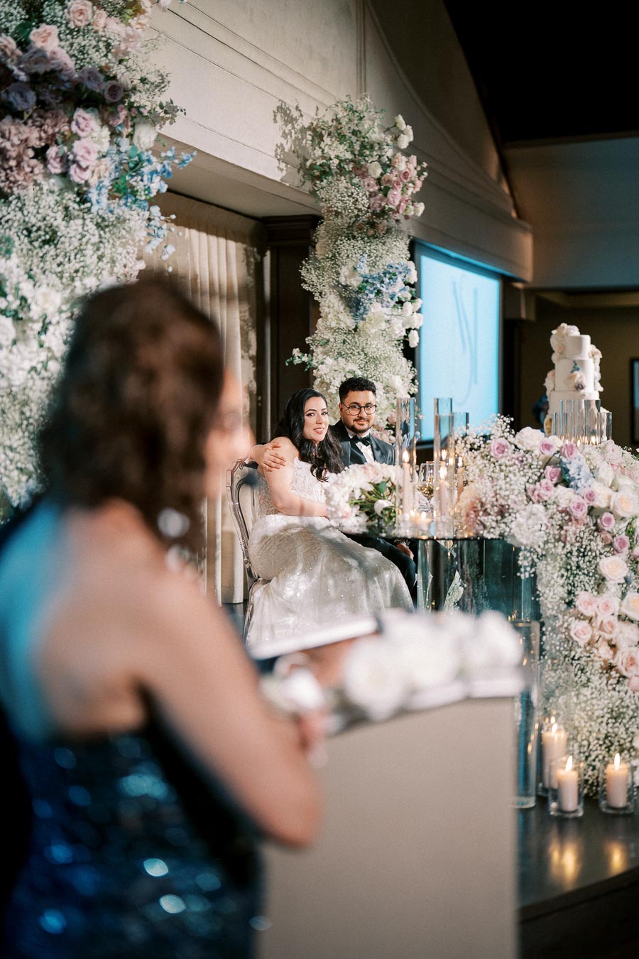 A bride and groom seated at a beautifully decorated wedding reception table, surrounded by lush floral arrangements and candles, listening to a speech with soft ambient lighting.
