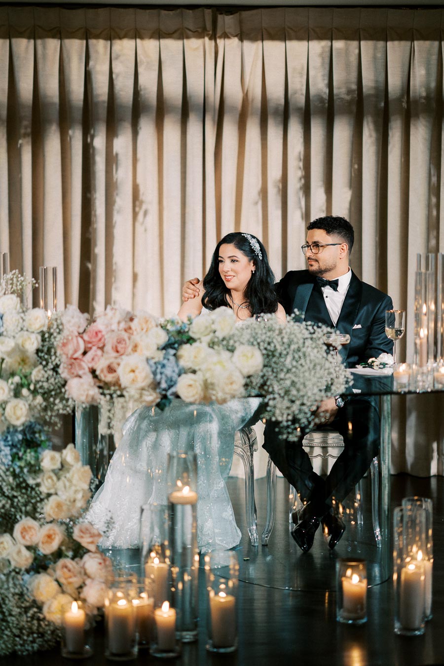 Elegant wedding couple seated at a table adorned with beautiful floral arrangements and surrounded by lit candles, creating a romantic atmosphere in a softly lit venue.
