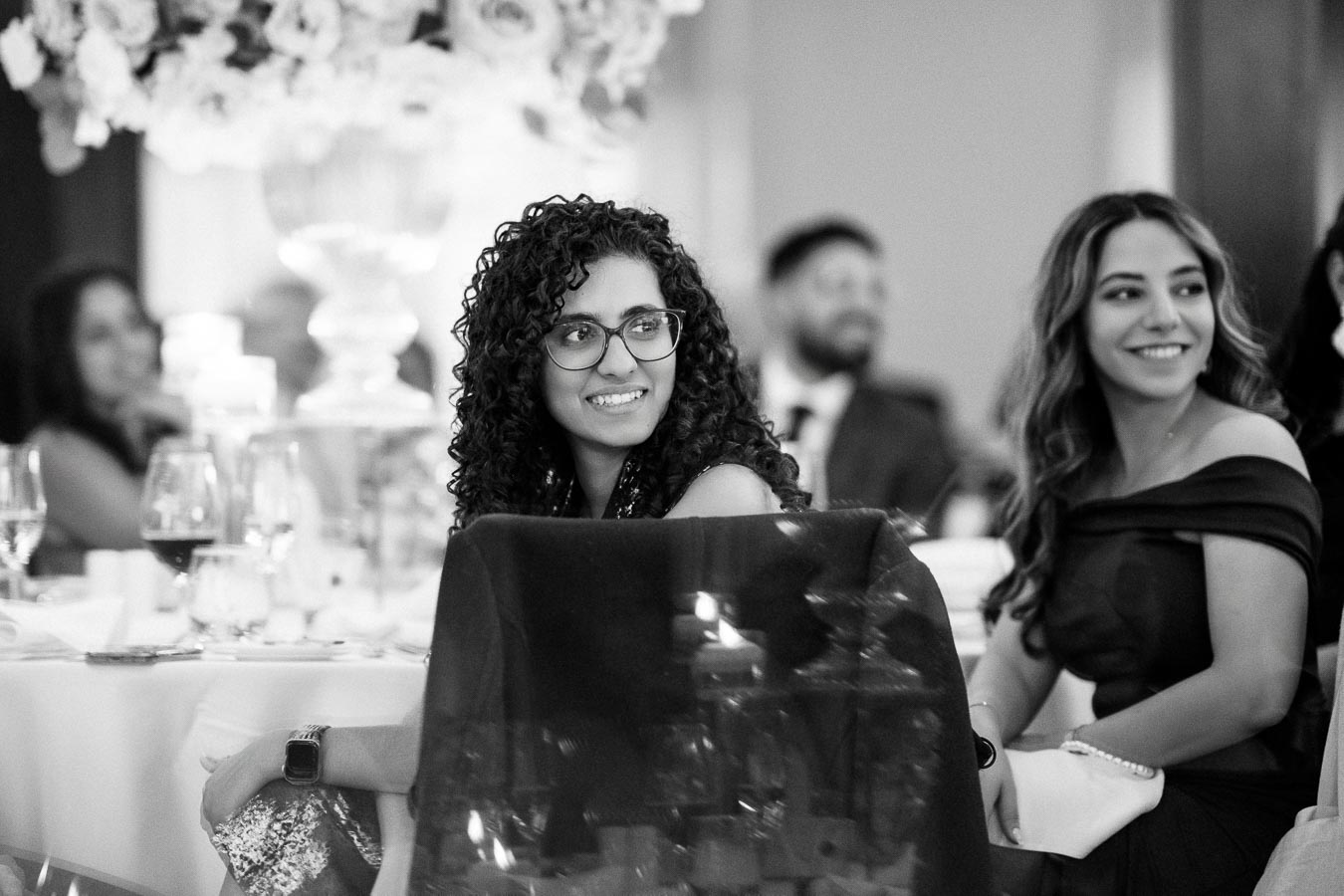 A black and white photo of people sitting at a formal event, smiling and engaged, with elegantly set tables and floral decorations in the background.