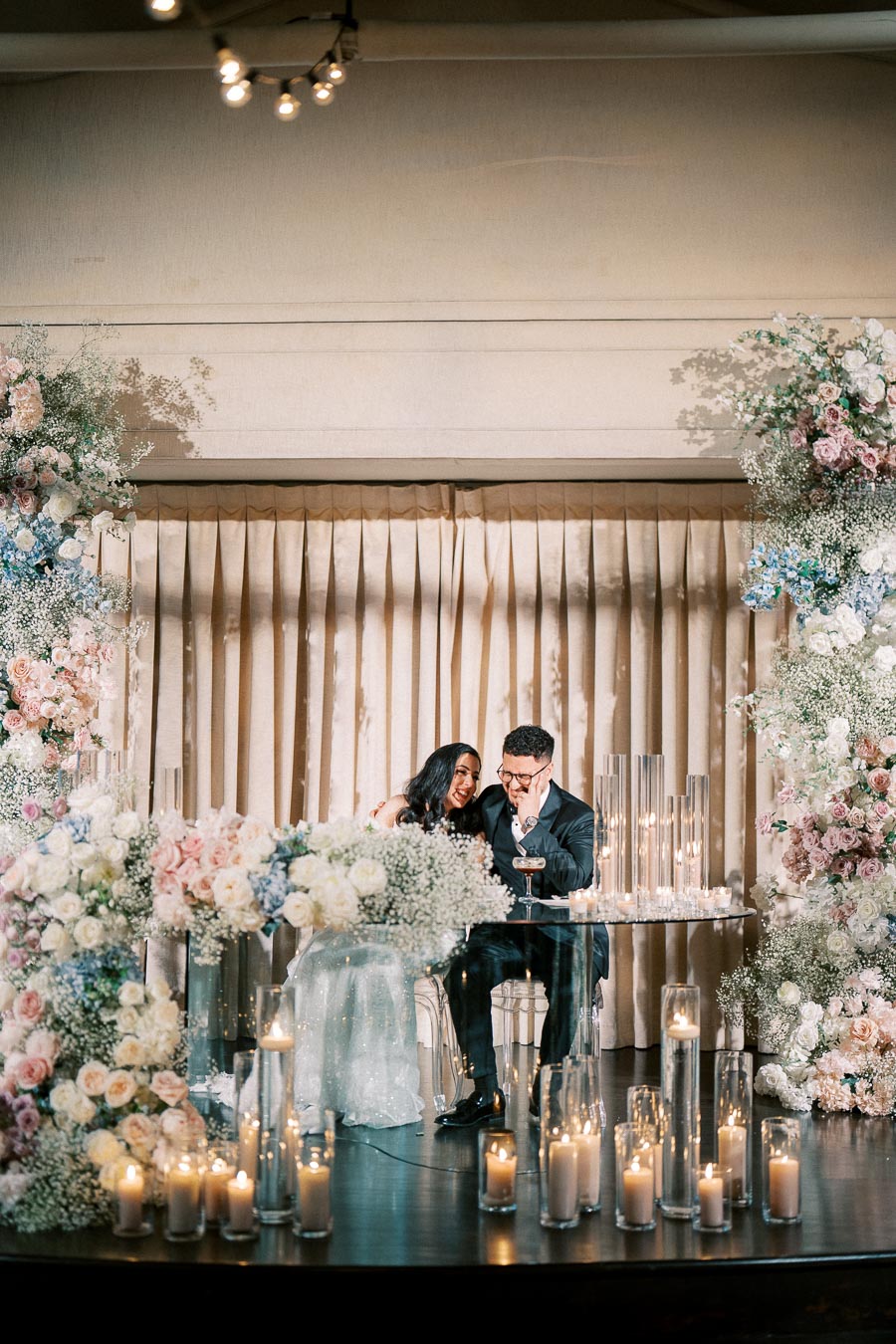 Elegant wedding reception scene with a couple sitting closely at a table adorned with floral arrangements and candles, surrounded by lush decorations of white, pink, and blue flowers, creating a romantic and intimate atmosphere.